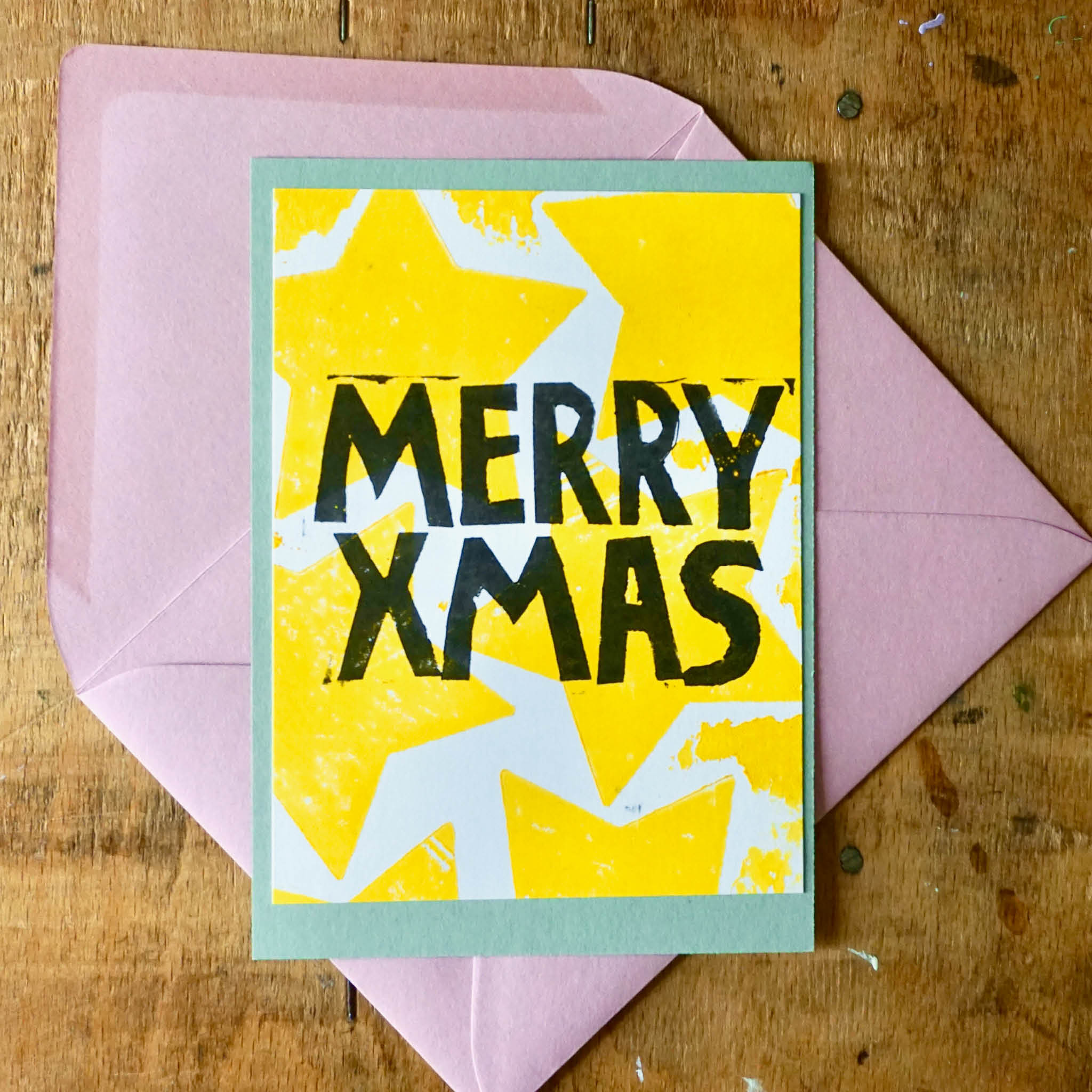 von Hand gedruckte Weihnachtskarte: Rosa Umschlag, grüne Karte als Basis, Weiße arte mit aufgedruckten gelben Sternen und Schriftzug MERRY XMAS